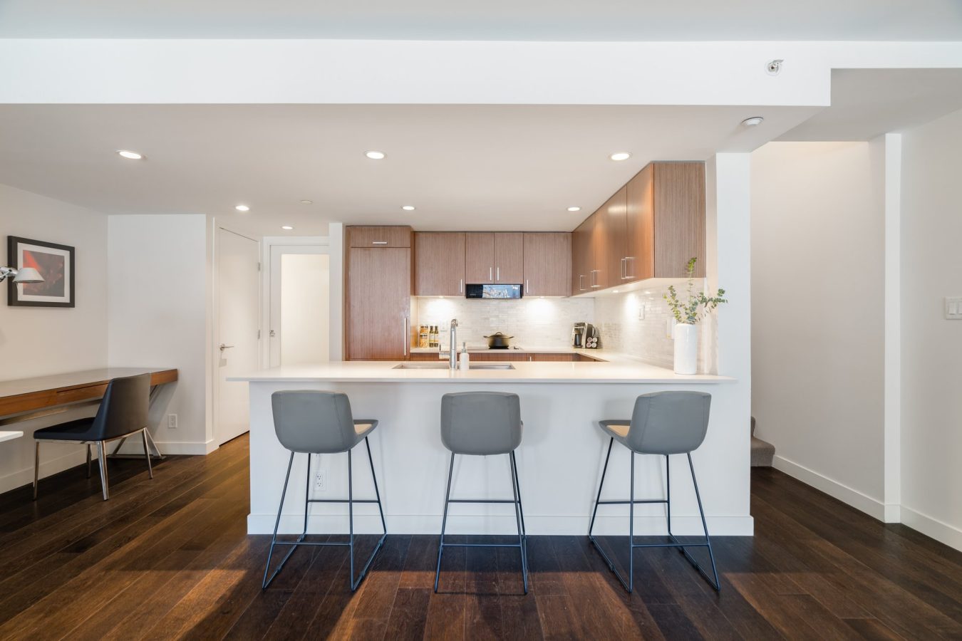 Modern kitchen with wood cabinets, white countertops, three gray bar stools, dark wood flooring, and built-in appliances. A small desk area with a chair is visible to the left, and a staircase is to the right.