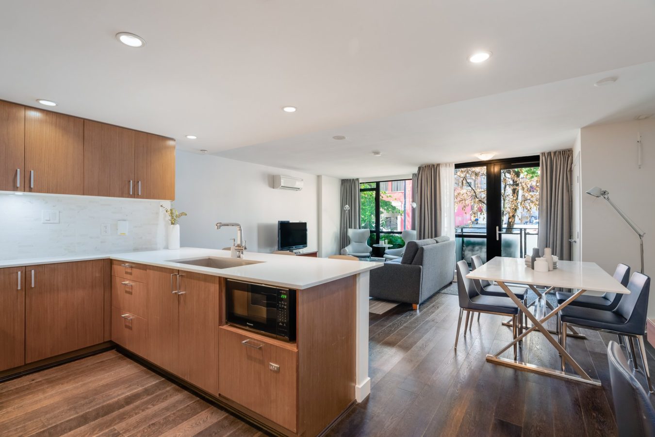 Modern open-plan kitchen and living area with wooden cabinets, built-in microwave, dining table with four chairs, grey sofas, TV, large windows, and glass door leading to a patio with greenery outside.
