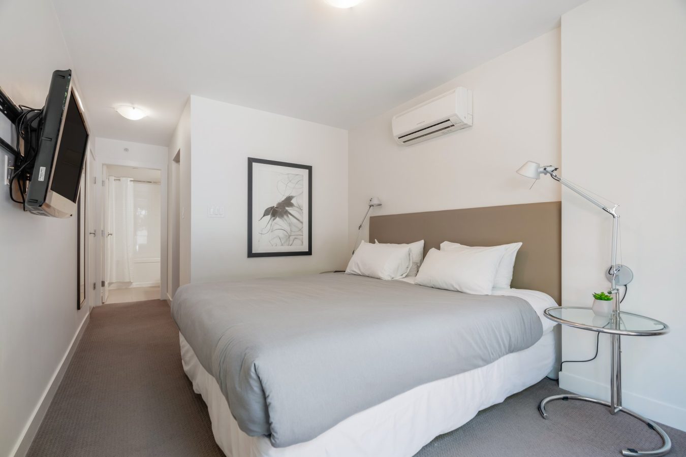 Minimalist bedroom with a large bed, gray bedding, two wall-mounted reading lamps, a round side table with a small plant, a wall-mounted TV, framed art, and white walls and carpet.
