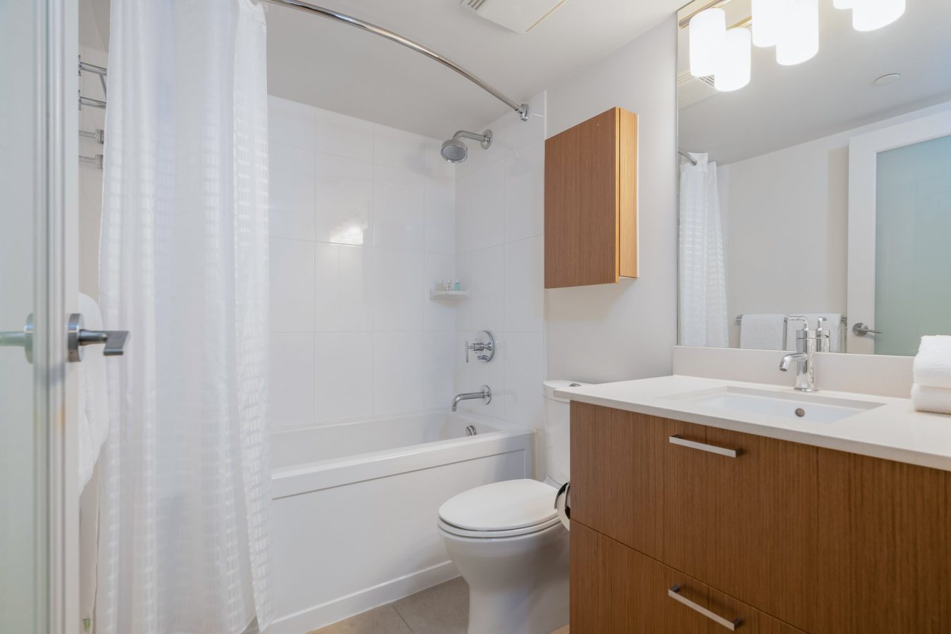 Modern bathroom with a white bathtub and shower, white curtain, wooden vanity with sink, wall-mounted cabinet, toilet, and bright overhead lighting. The space is clean and minimalistic.