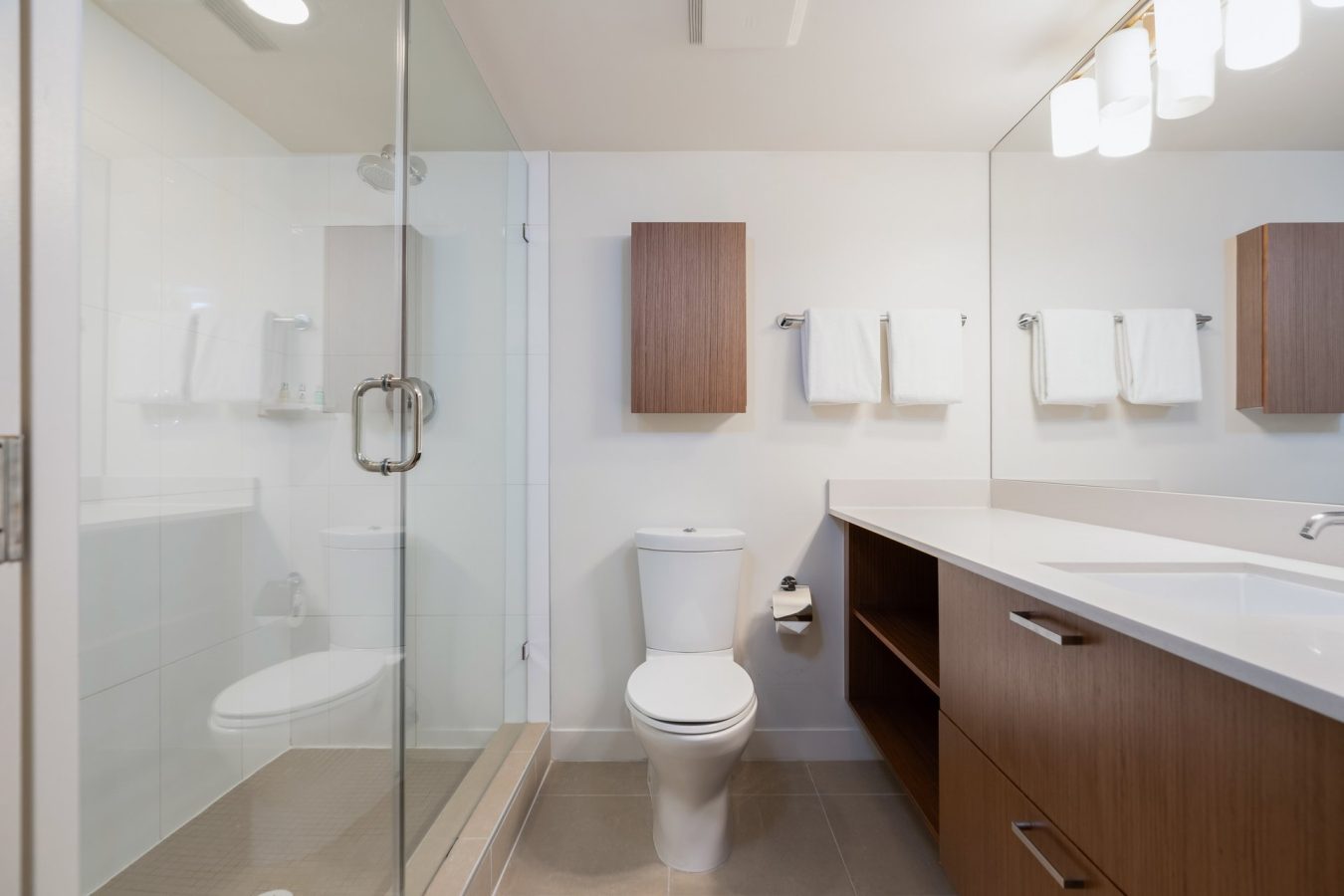Modern bathroom with a glass-enclosed shower, a white toilet, wooden vanity with sink, large mirror, and towels hanging on the wall. The room features neutral colors and clean, minimalist design.
