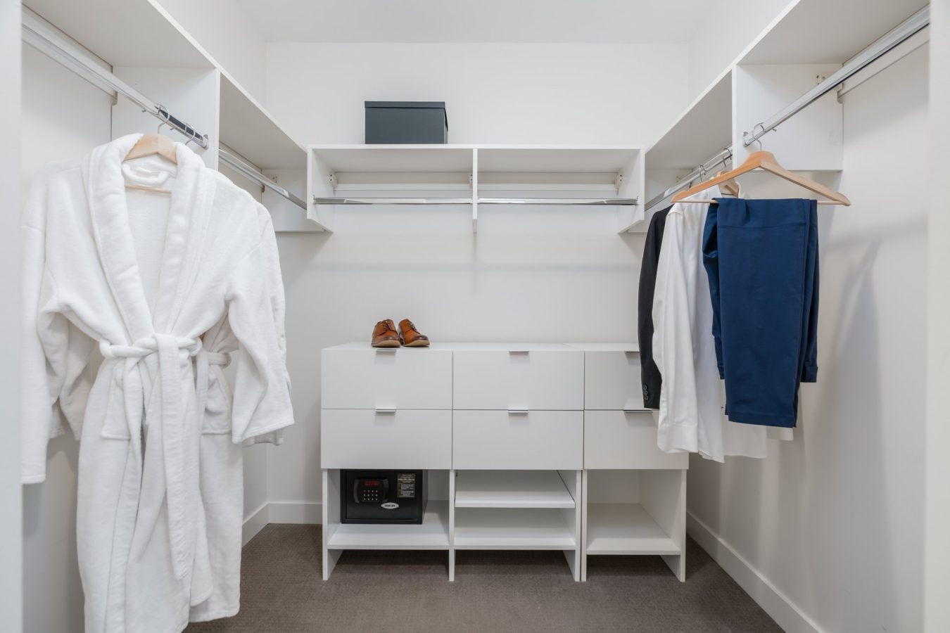 A modern, white walk-in closet with shelves, a safe, a robe hanging on the left, a pair of shoes on the middle shelf, and several neatly hung clothes on the right. The space is clean and organized.