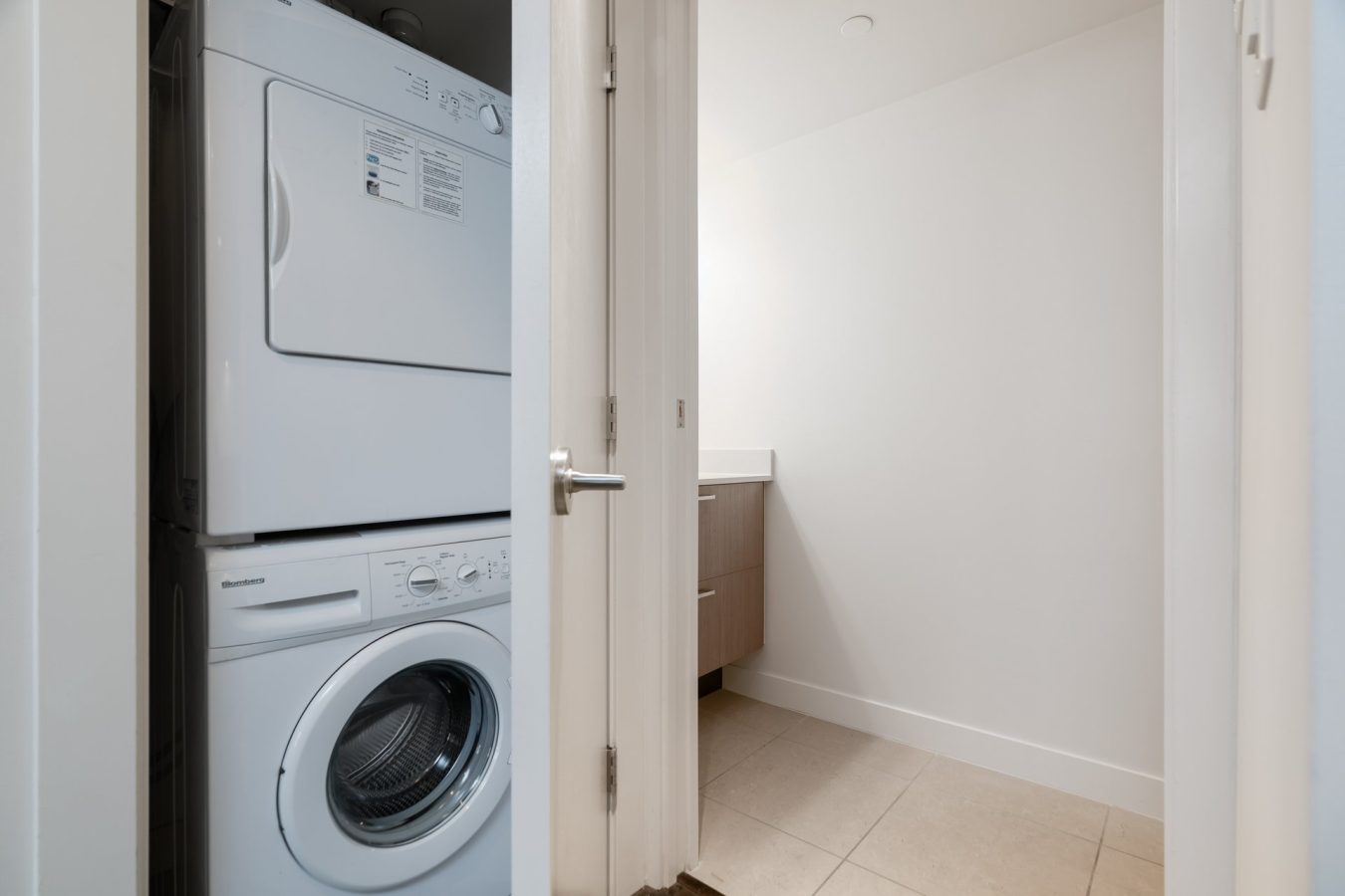 A small laundry area with a stacked washer and dryer unit next to an open door, leading into a brightly lit room with a tiled floor and a wooden cabinet.