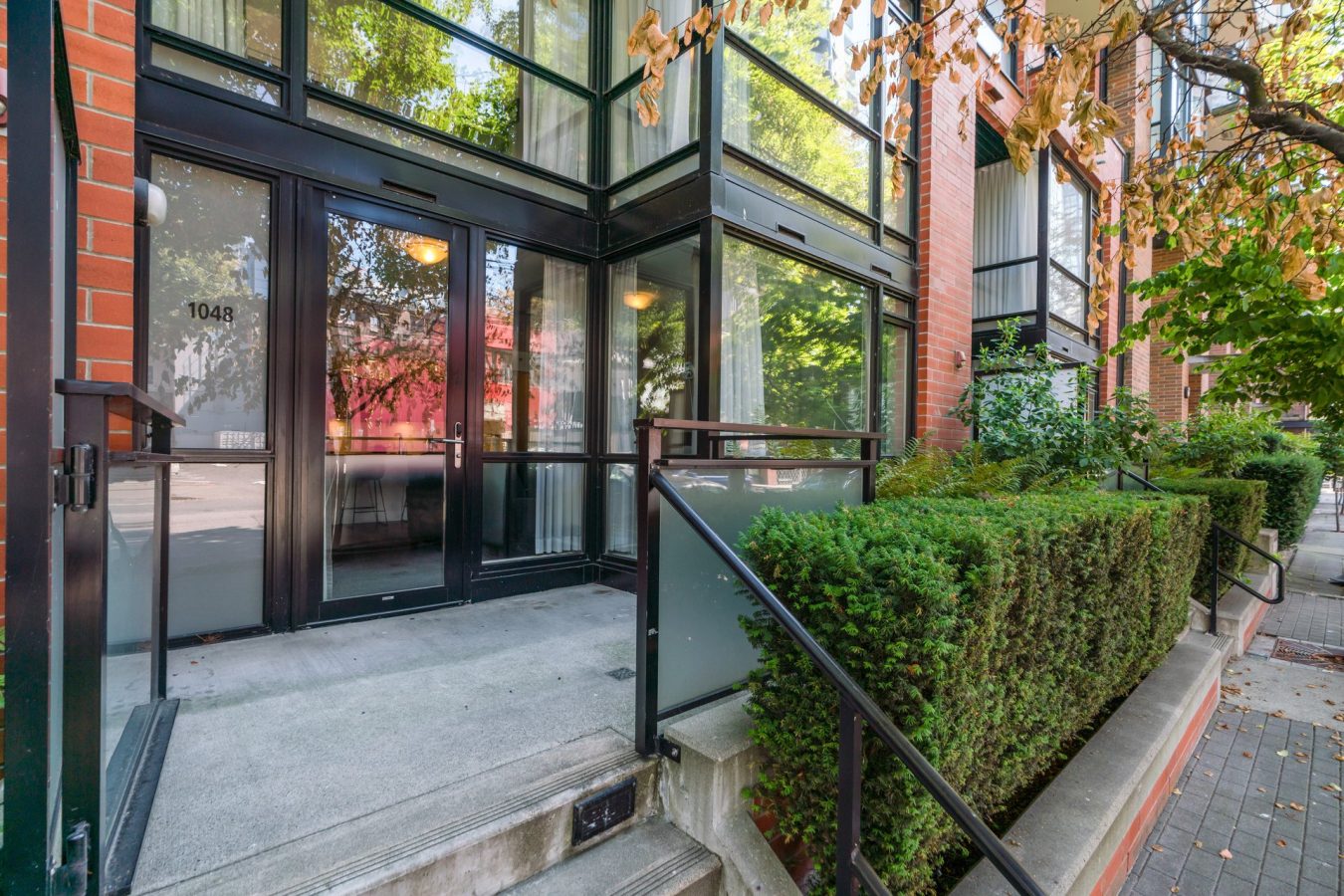 Glass entrance of a modern brick building with black metal frames, a small concrete staircase, hedge-lined walkway, and trees with green leaves along the sidewalk. Address number 1048 is visible on a door.