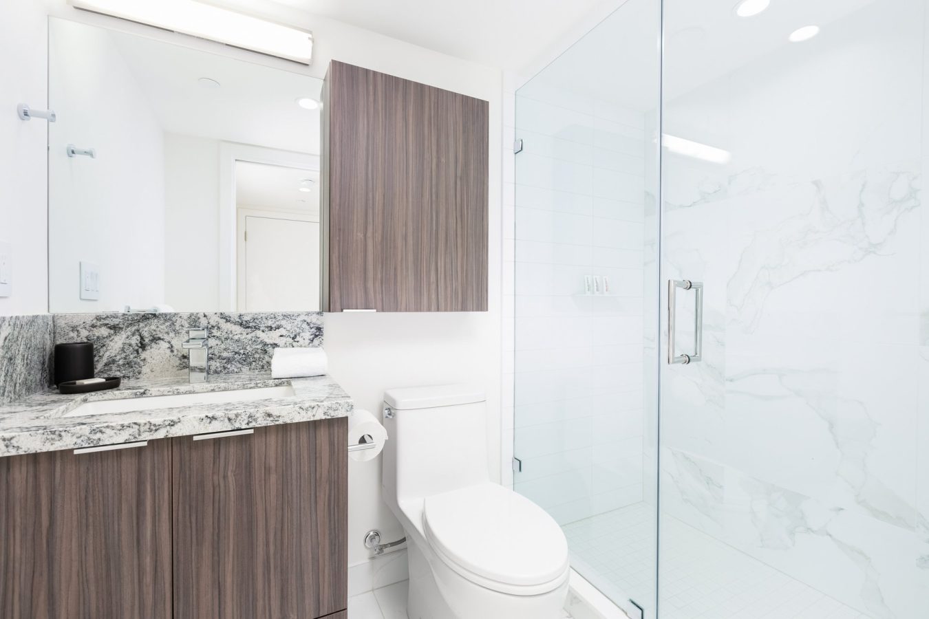 Modern bathroom with a wood vanity, marble countertop, wall-mounted cabinet, toilet, and glass-enclosed shower with white marble tiles. The space is bright and clean with white walls and floor.