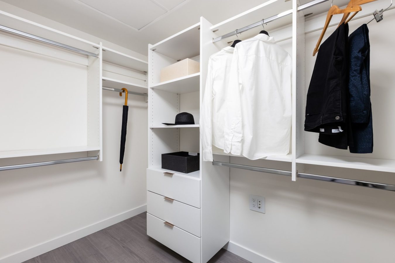 A modern, organized walk-in closet with white shelves, hanging rails, neatly hung shirts and jackets, an umbrella, a hat, storage boxes, and a set of drawers on a light wood floor.