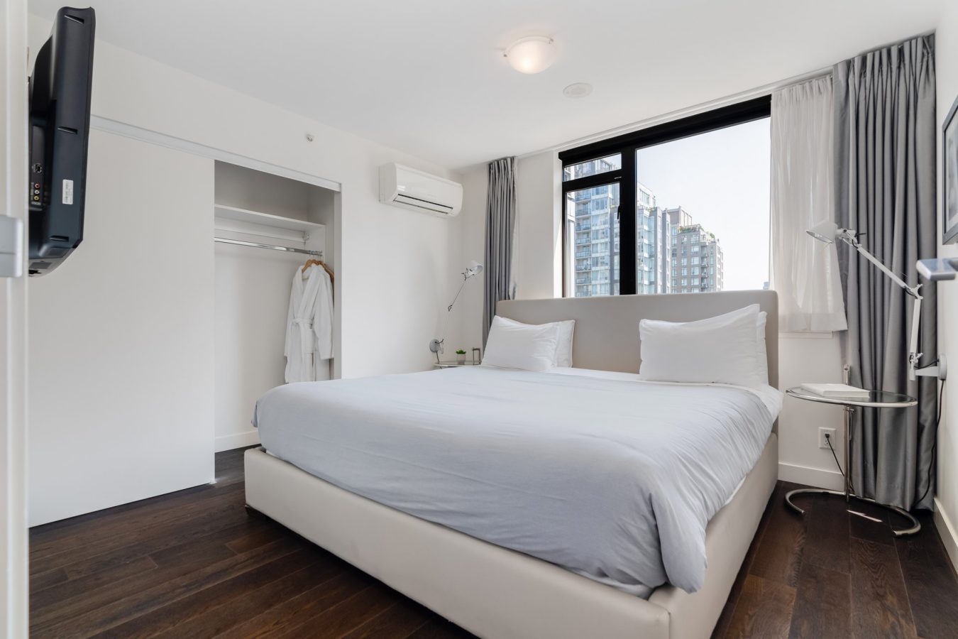 A modern, minimal bedroom with a large bed, white linens, wooden floor, open closet with a white robe, large window with city view, and light gray curtains. A wall-mounted TV and desk lamp are also visible.