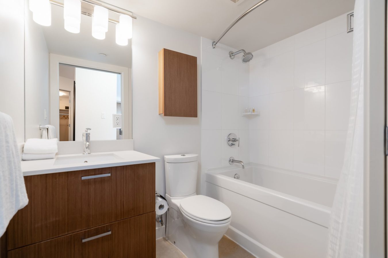 A modern bathroom with a wooden vanity, white sink, toilet, wall cabinet, and a bathtub-shower combo with white tiles. A large mirror and bright lights are above the sink. Towels are neatly placed.