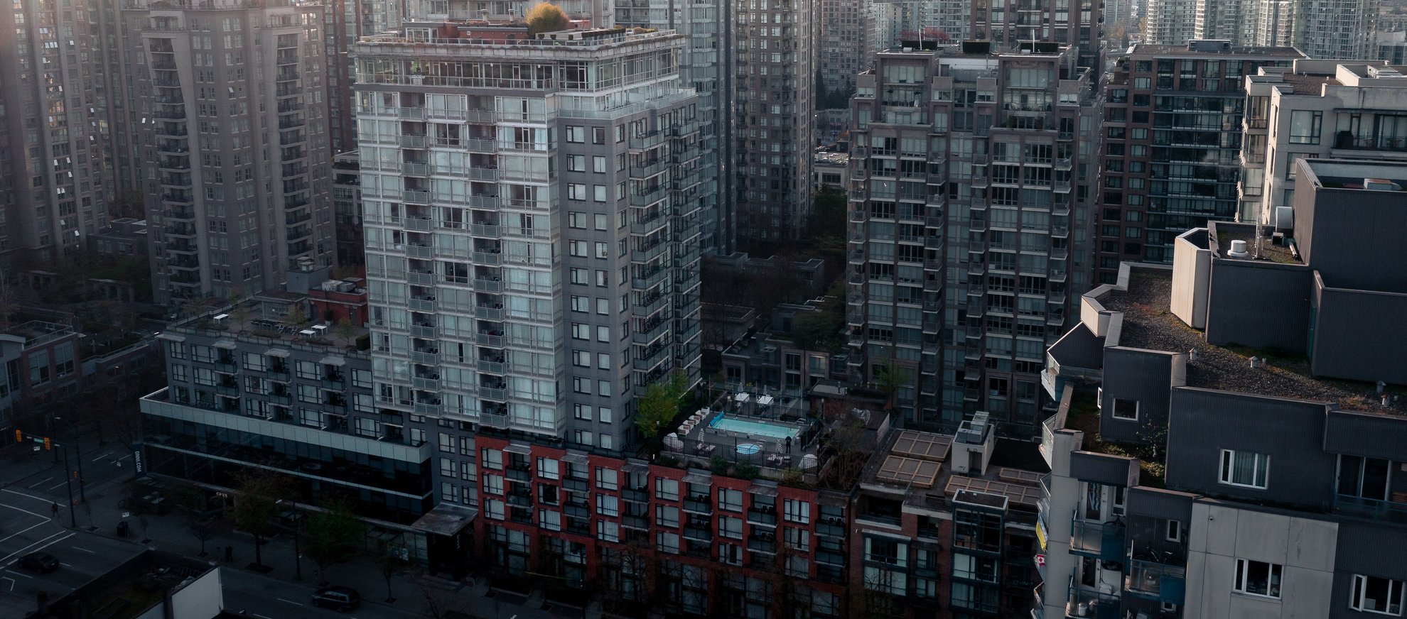 A cluster of modern high-rise apartment buildings in a city; one building features a rooftop pool, while streets and trees are visible below. The scene appears overcast and densely populated.