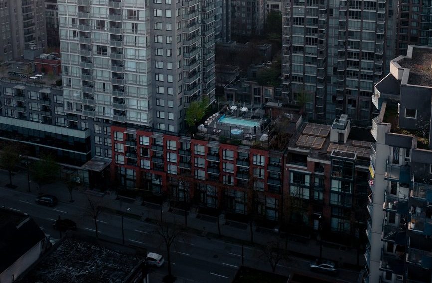 A rooftop swimming pool is surrounded by lounge chairs atop a mid-rise building, with tall modern apartment buildings and a quiet street visible below at dusk.
