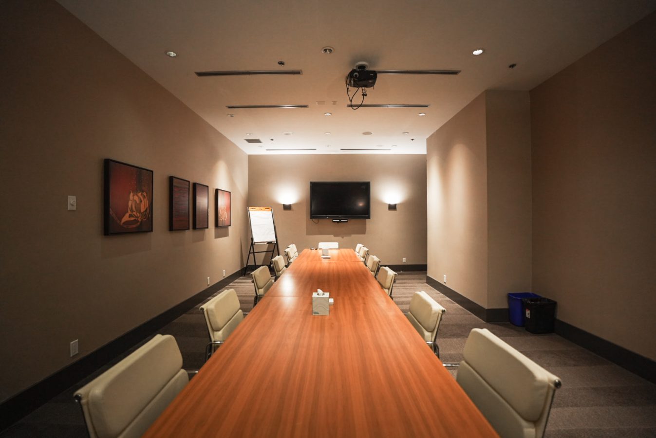 A modern conference room with a long wooden table surrounded by white chairs, a TV on the wall, a flip chart, framed artwork, and ceiling lights, creating a professional meeting environment.