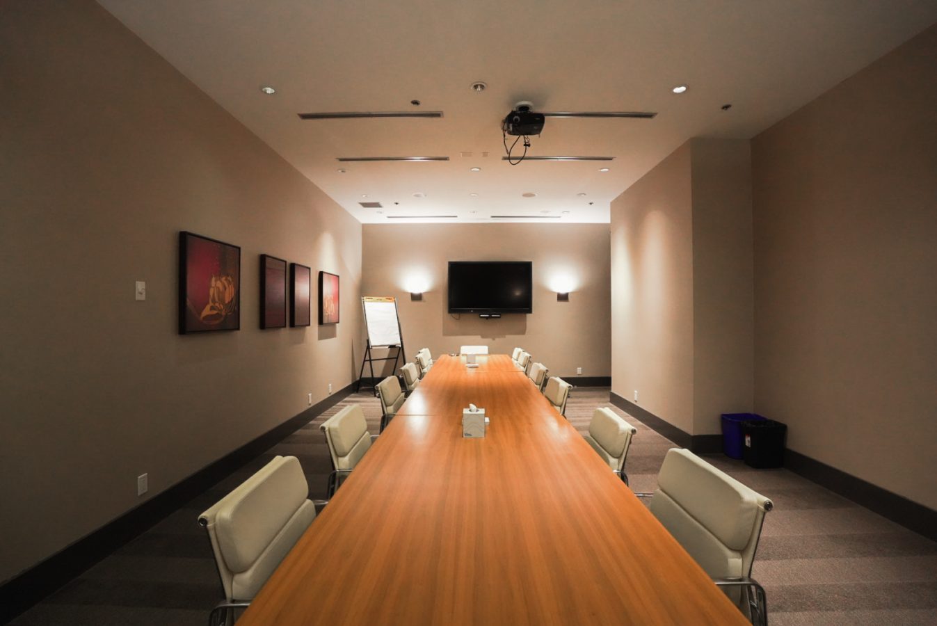 A modern conference room with a long wooden table surrounded by white chairs, a wall-mounted TV, a projector on the ceiling, framed artwork on the wall, and a flip chart in the corner. The room is well-lit.