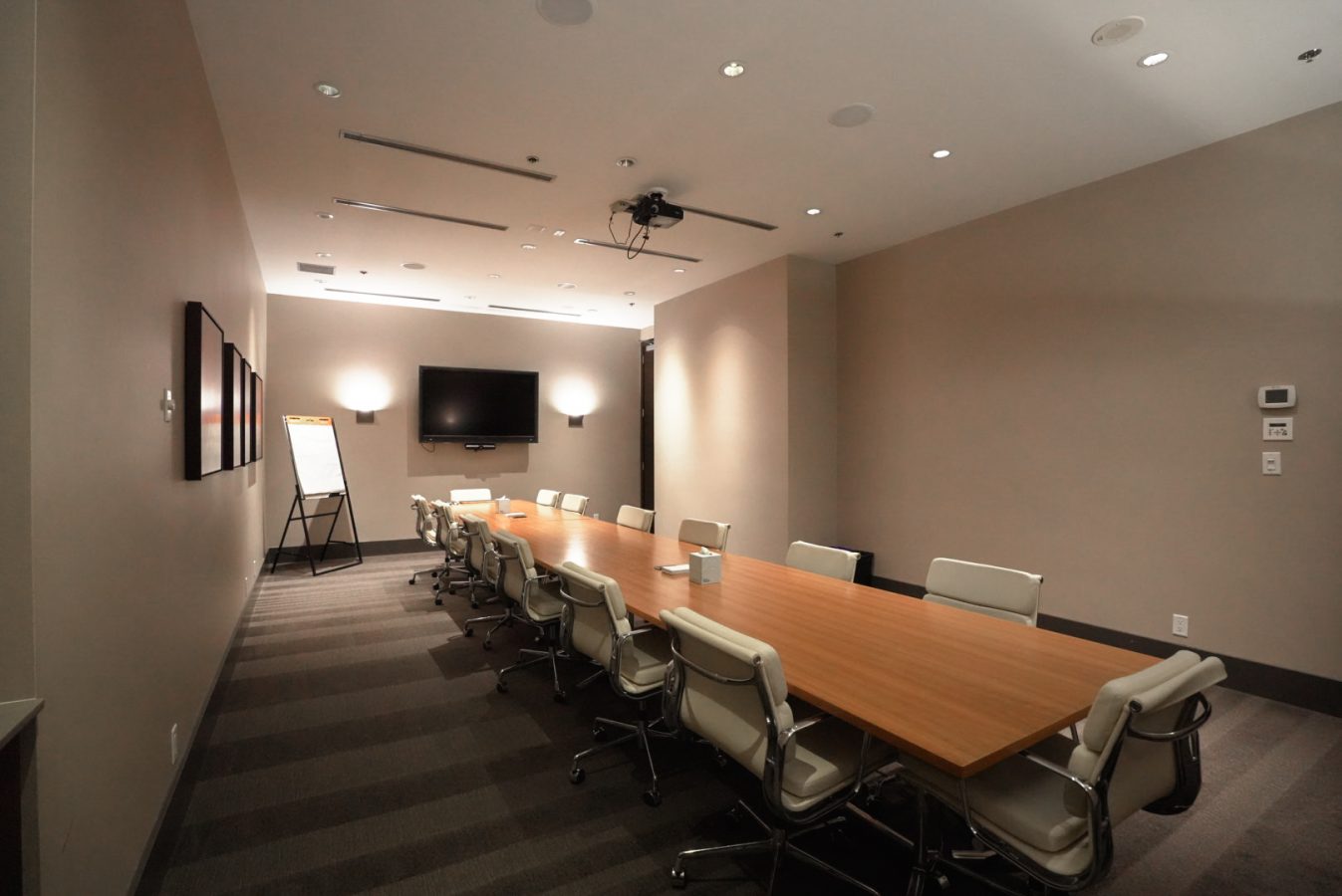 A modern conference room with a long wooden table, white office chairs, a wall-mounted TV, a flip chart, and neutral-colored walls with framed art. The room is well-lit and has a minimalist design.