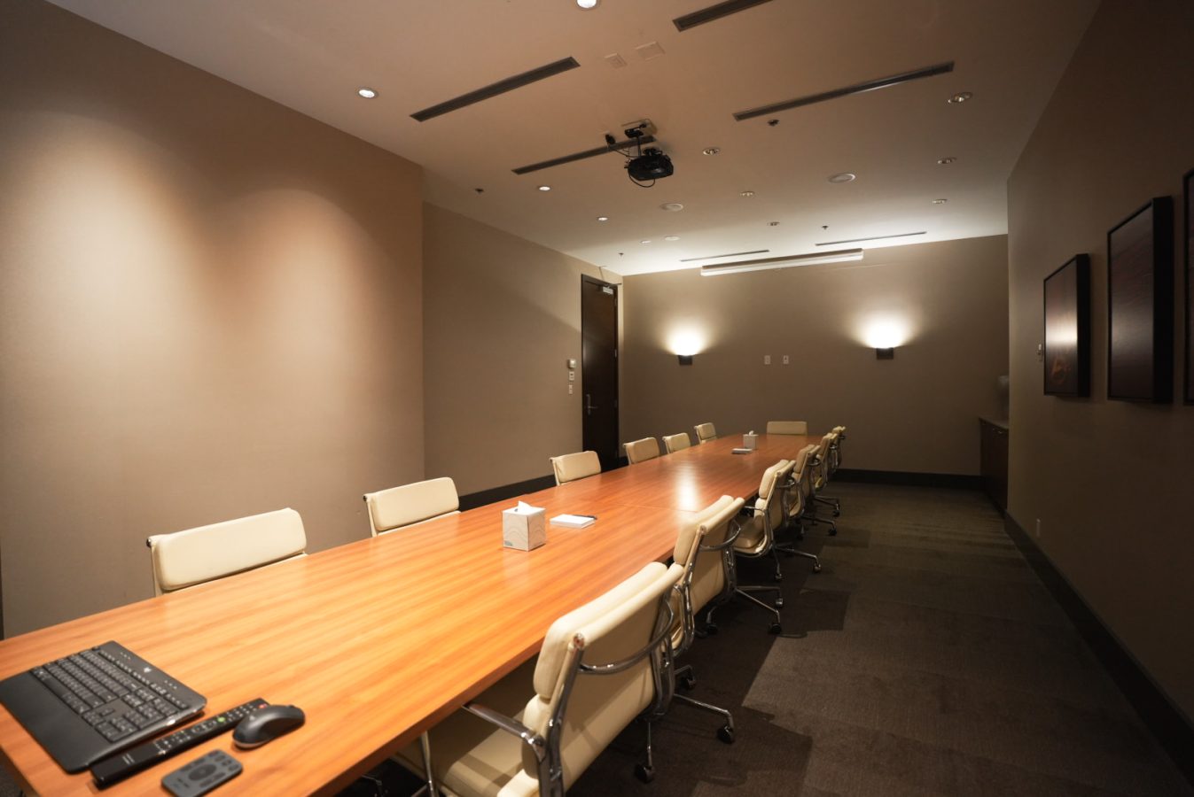 A modern, empty conference room with a long wooden table, cream swivel chairs, a wall-mounted screen, computer keyboard, mouse, ceiling projector, and neutral-colored walls with soft lighting.