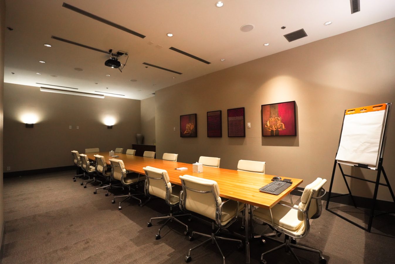 A modern conference room with a long wooden table, white chairs, a projector on the ceiling, three framed artworks on the wall, and a flip chart with blank paper in the corner. The room is brightly lit.