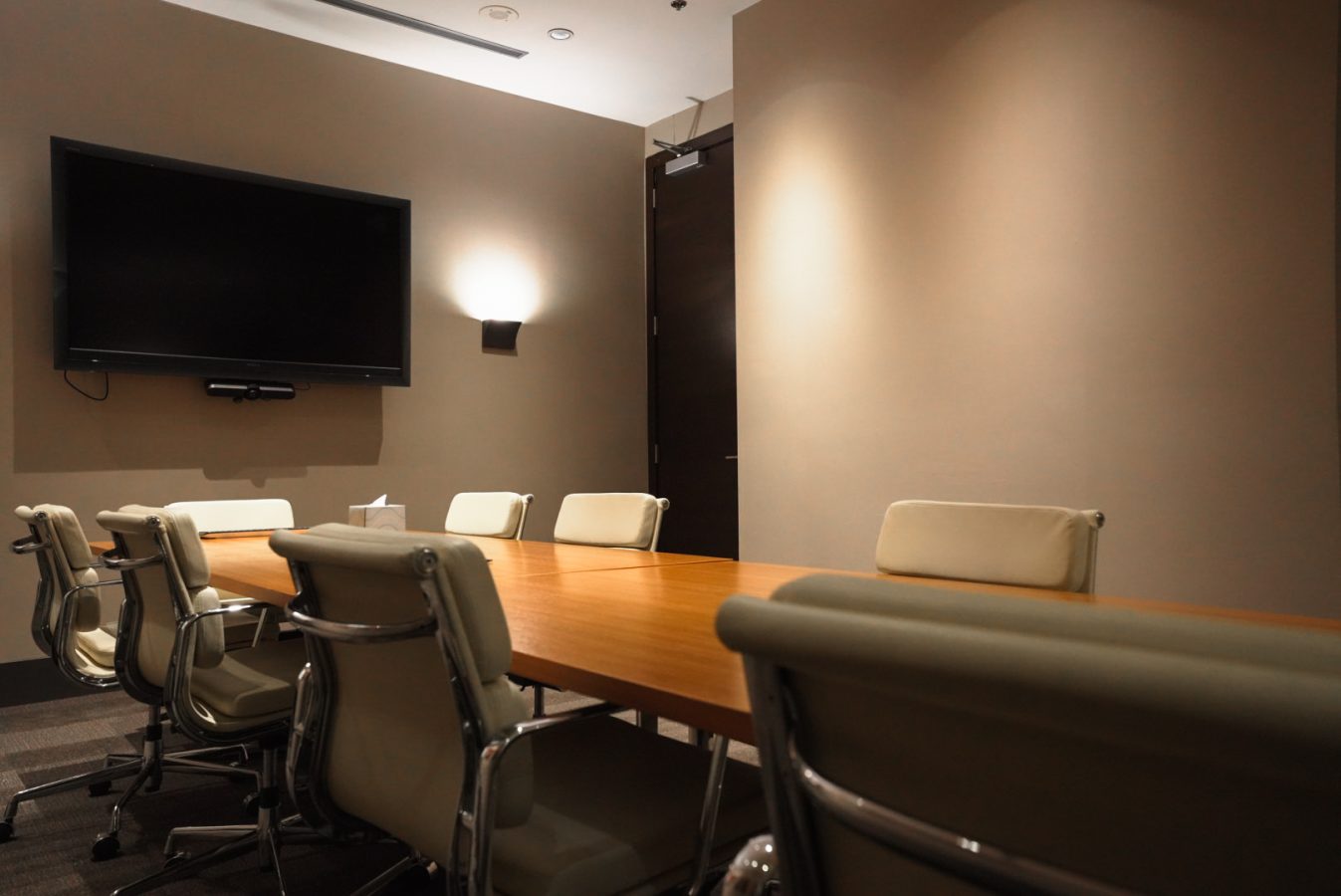 A modern conference room with a wooden table surrounded by beige chairs, a large flat-screen TV mounted on the wall, soft lighting, and a closed dark wooden door in the background.