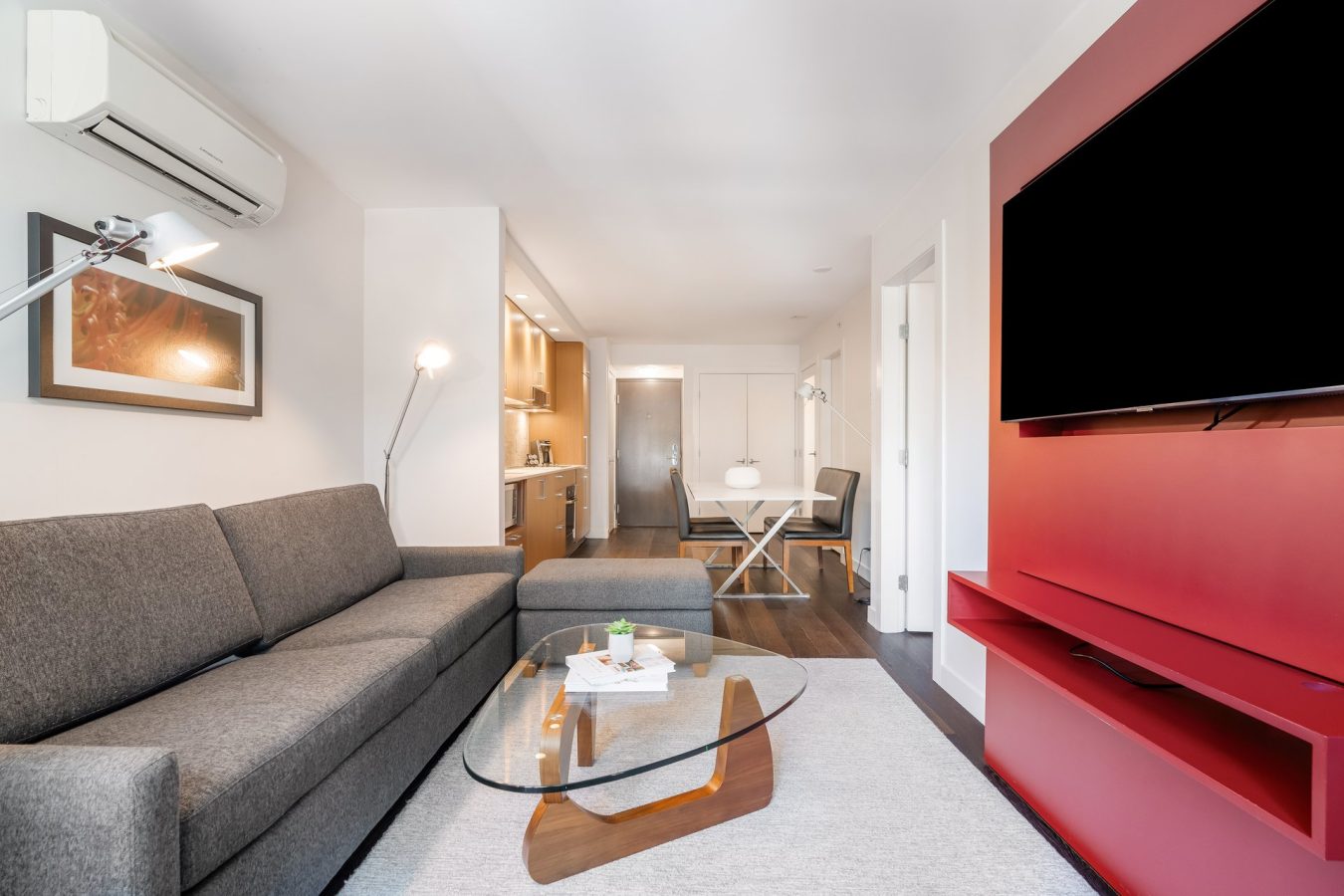 Modern living room with a gray sectional sofa, glass coffee table, wall-mounted TV on a red feature wall, kitchen area in the background, and bright natural lighting.