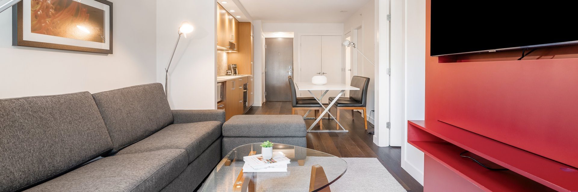 Modern apartment living room with a gray sofa, glass coffee table, wall-mounted TV on a red accent wall, and a dining area with a white table and two black chairs. Kitchen area and hallway visible in the background.