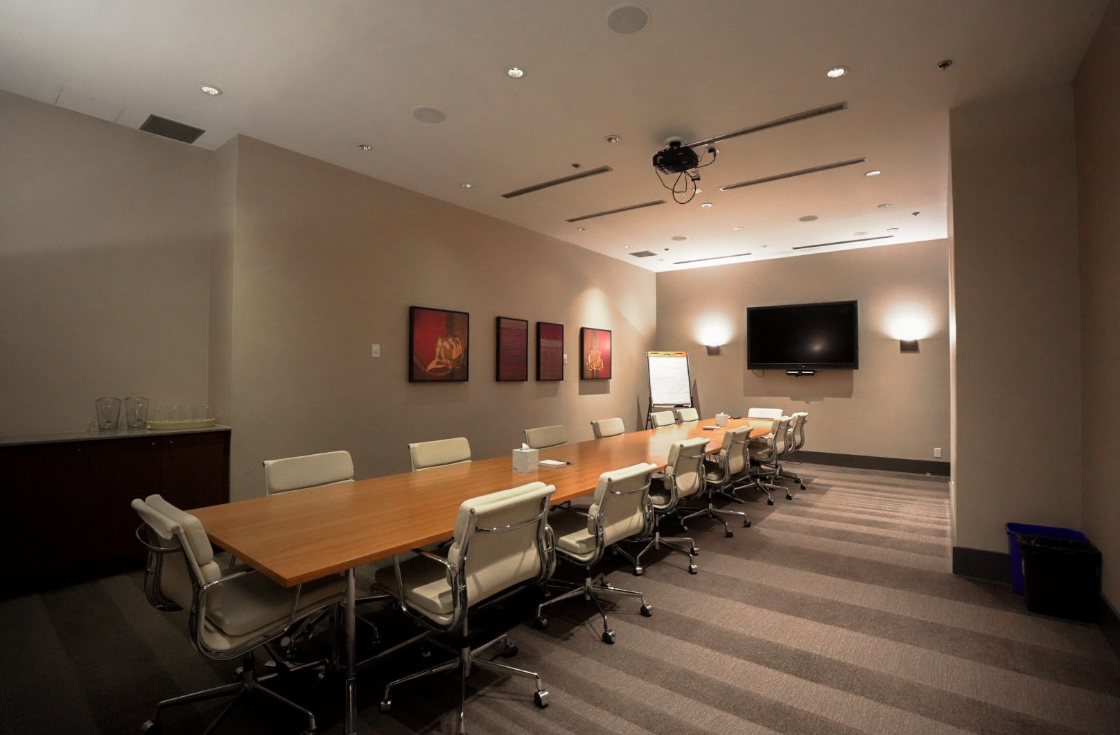 A modern conference room with a long wooden table surrounded by white chairs, a wall-mounted TV, a flip chart, abstract artwork on the wall, and soft overhead lighting.