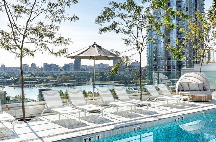 Rooftop pool with white lounge chairs, umbrella, and a round daybed. Trees provide shade, and tall city buildings and a river are visible in the background under a clear sky.