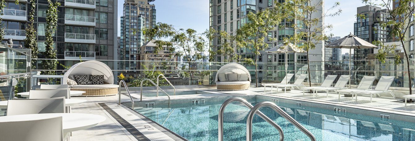 Modern rooftop pool surrounded by lounge chairs, daybeds, and umbrellas, with glass railings and tall buildings in the background, under a bright, clear sky.