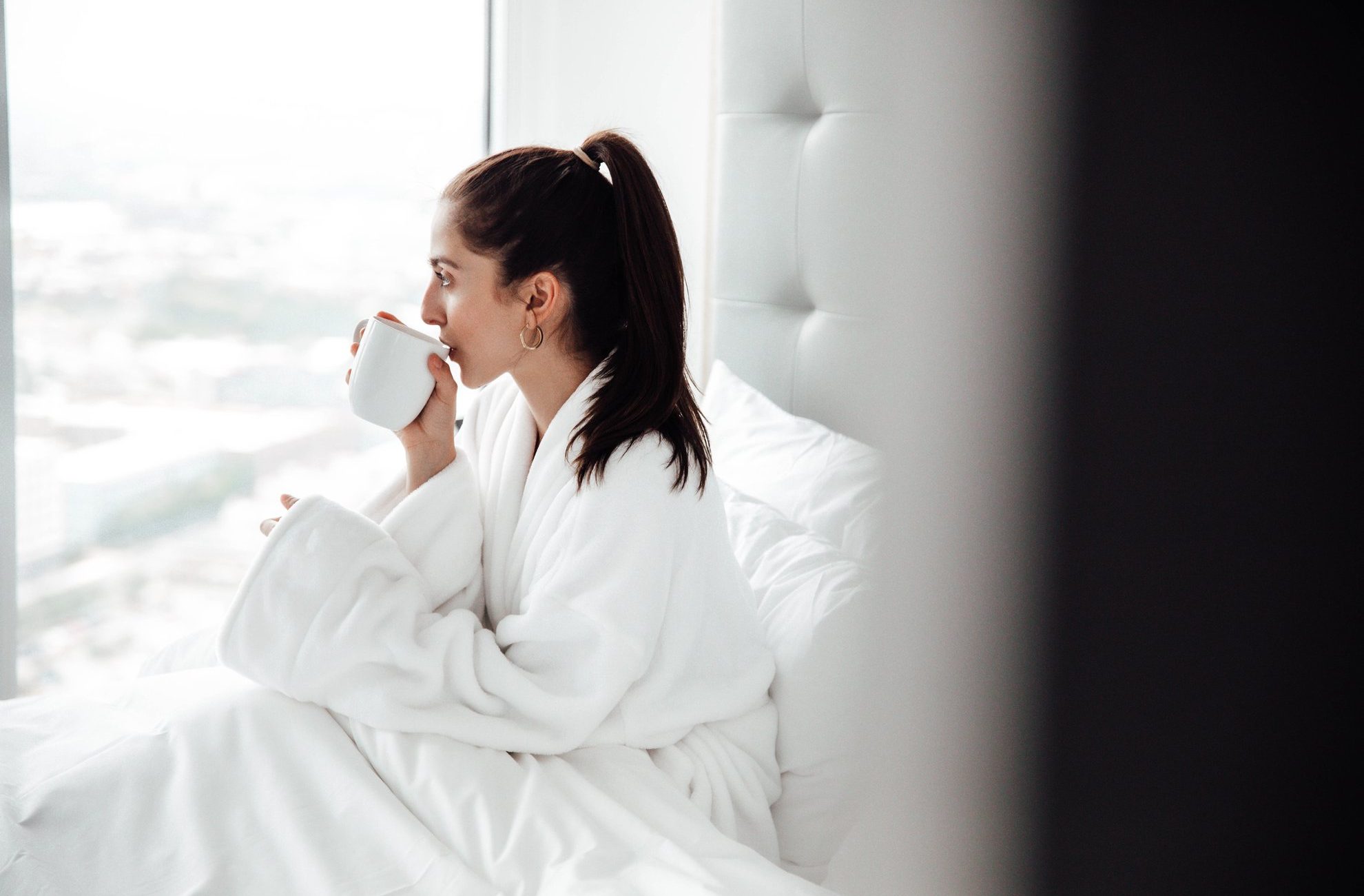 A woman in a white bathrobe sits on a bed by a large window, sipping from a white mug. The room is bright with natural light, and she appears relaxed and contemplative.
