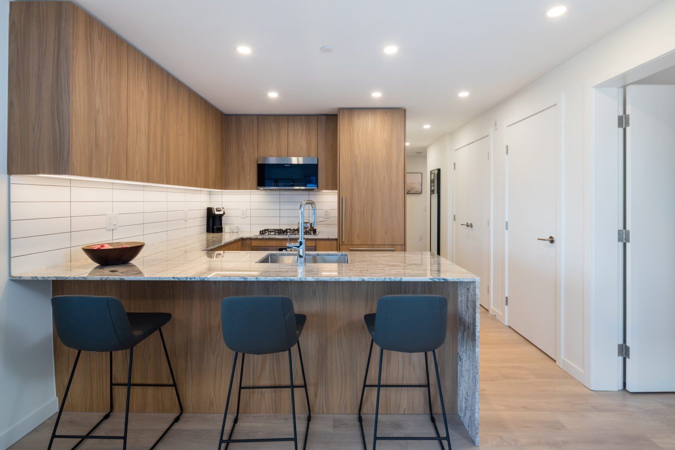 Modern kitchen with wood cabinets, white tile backsplash, a marble island with three black bar stools, built-in appliances, and recessed ceiling lights. The room has light wood flooring and white walls.