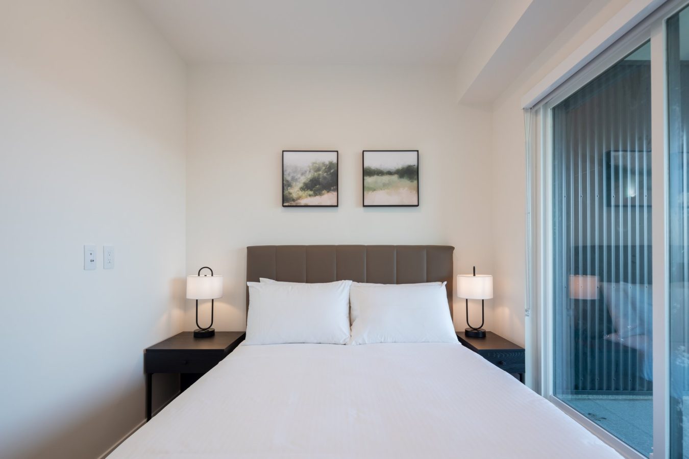A neatly made bed with white linens is centered between two nightstands, each with a lamp. Two landscape paintings hang above the bed, and a sliding glass door is visible on the right. The room has a clean, minimalist style.