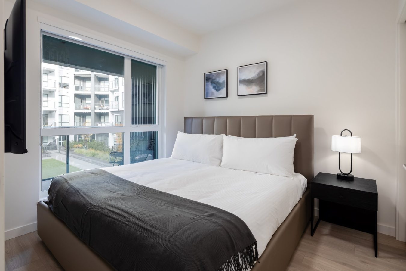 A modern bedroom with a large bed, white bedding, gray headboard, black throw blanket, black nightstand with a lamp, two framed pictures above the bed, and a window overlooking an apartment complex.