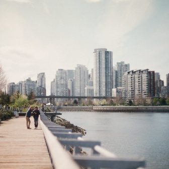 Two people walk together on a wooden boardwalk along the water, with modern high-rise buildings and greenery in the background under a partly cloudy sky.
