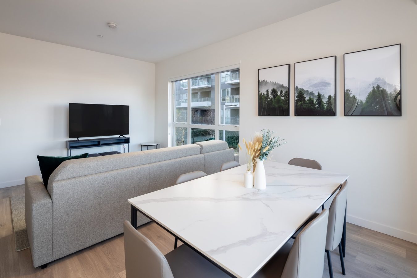 A modern living and dining area with a gray sofa facing a TV, a marble dining table with chairs, two vases with dried flowers, large window, and three framed nature prints on a white wall.