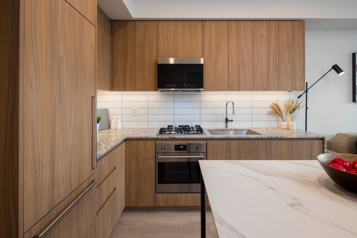 Modern kitchen with wood cabinetry, stainless steel appliances, white tile backsplash, and a marbled island countertop. A bowl of red apples sits on the island, and a sleek black lamp is visible in the background.