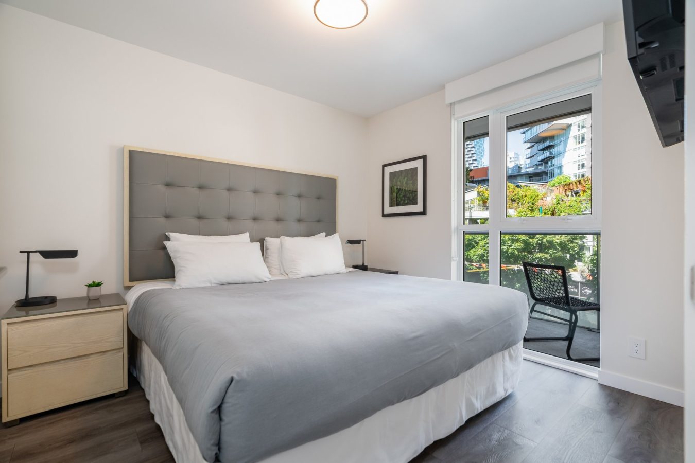 A modern bedroom with a large bed, gray tufted headboard, neutral bedding, two nightstands with lamps, a window with a view of greenery and buildings, and a chair on a small balcony.