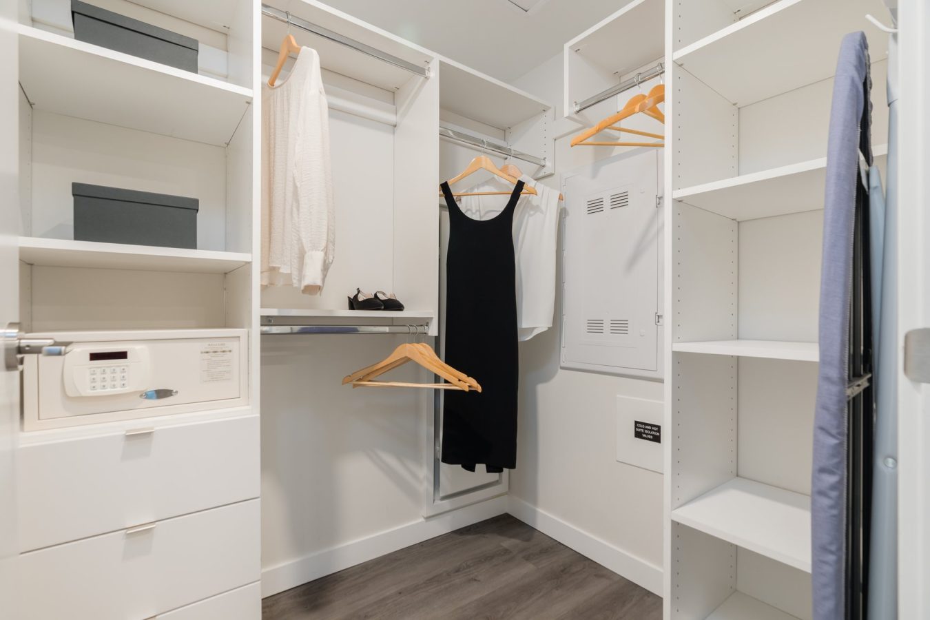 A modern walk-in closet with white shelves and drawers, a safe, a few hanging clothes including a white sweater and black dress, and some storage boxes. The floor is wood, and the space is well-organized and tidy.