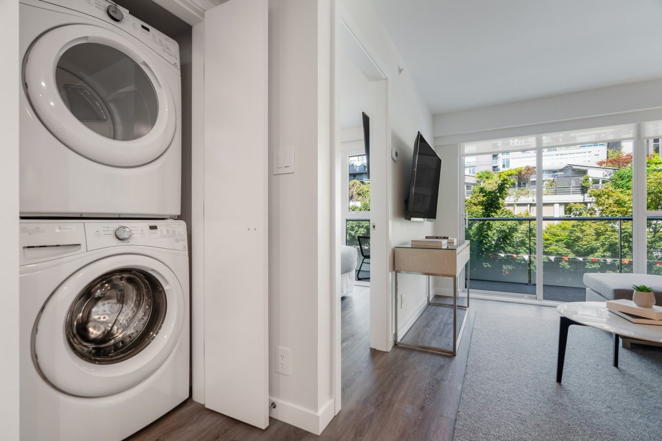 A modern apartment with a stacked washer and dryer in a small closet, an open living area with a TV, a desk, a sofa, and large windows overlooking greenery and city buildings.
