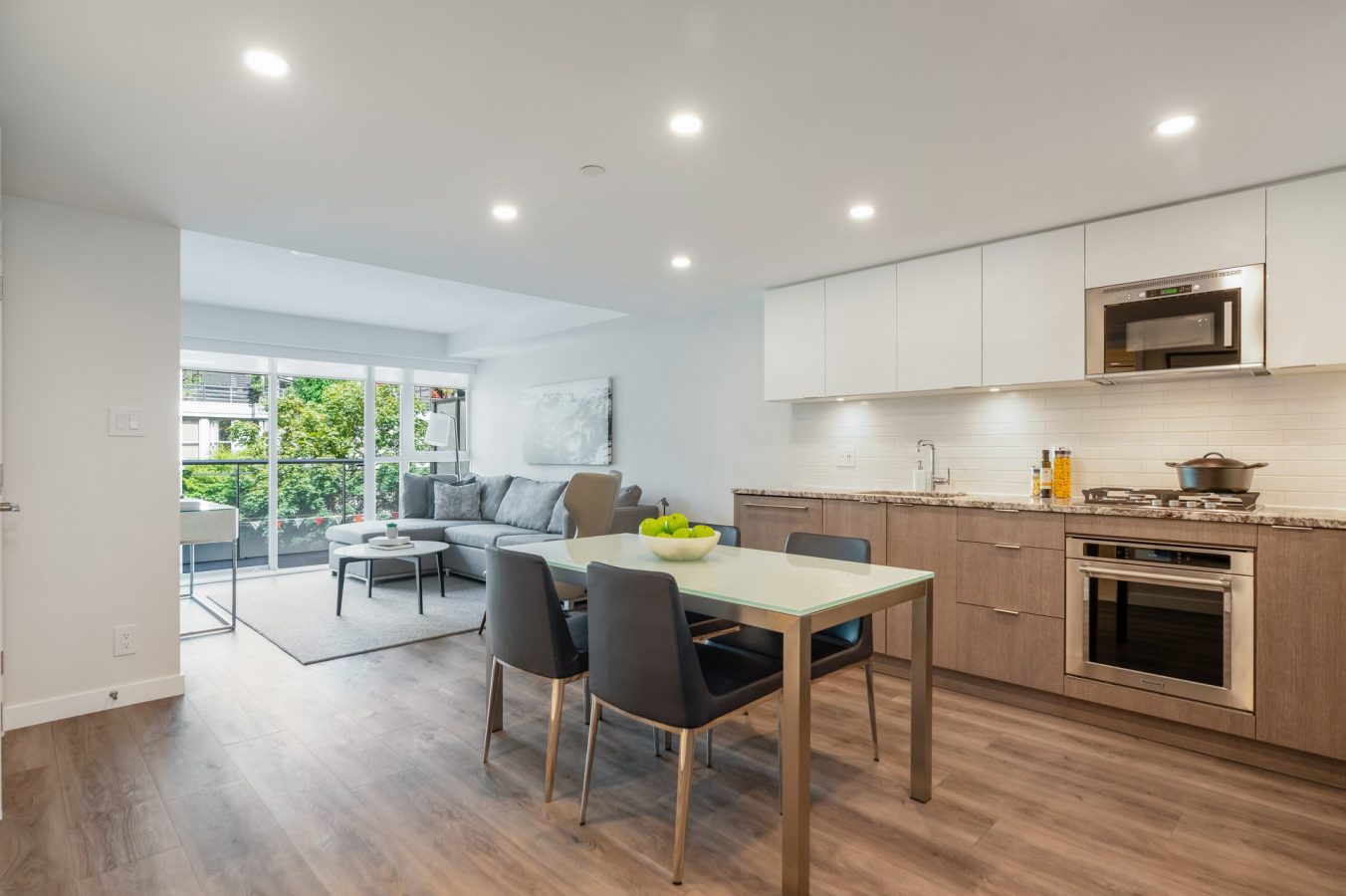 Modern open-plan kitchen and living area with wood flooring, dining table with four chairs, built-in appliances, gray sofa, artwork, and large windows leading to a balcony with greenery visible outside.