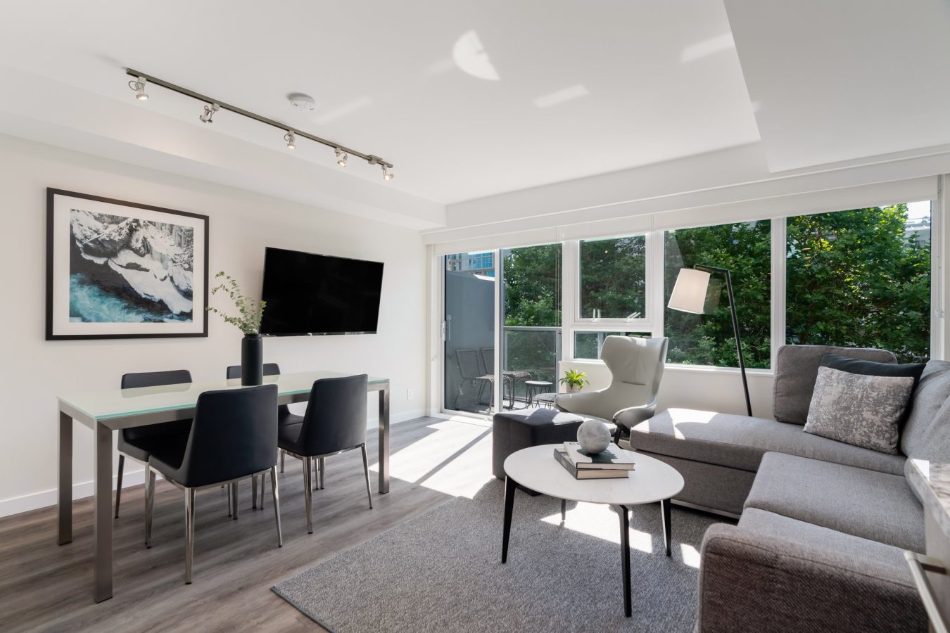 Modern living and dining area with gray sofa, round coffee table, wall-mounted TV, dining table with chairs, floor-to-ceiling windows, and balcony view of greenery. Bright and minimal decor.