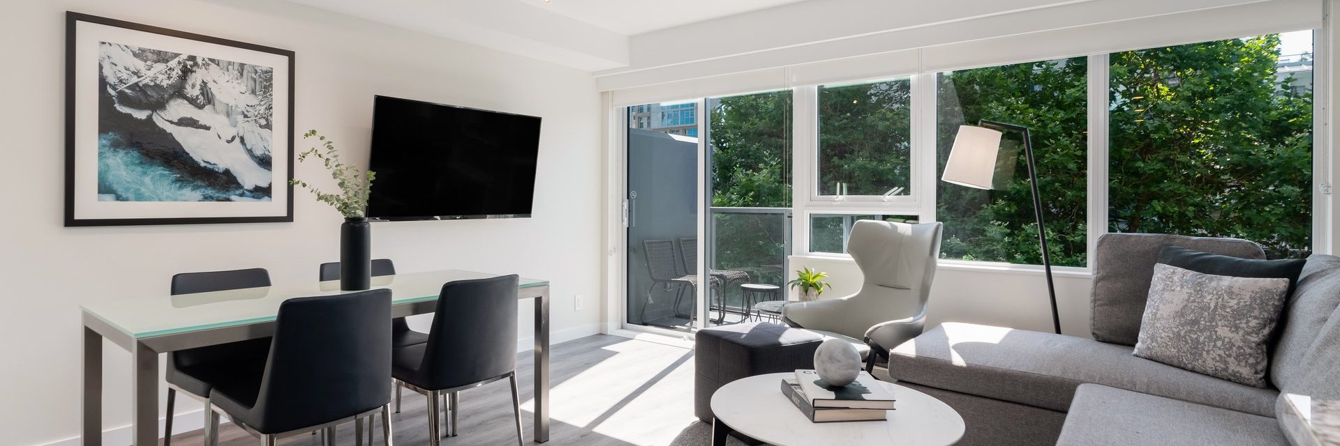 Modern living room with large windows, gray sectional sofa, armchair, wall-mounted TV, dining table with four chairs, and minimal decor. Sunlight streams in, highlighting greenery outside.