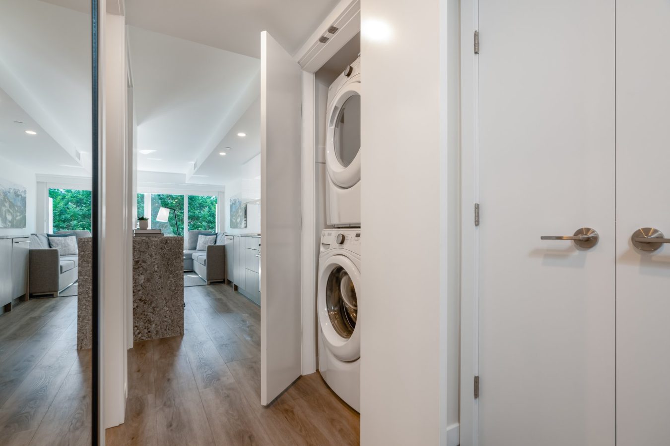 A stacked washer and dryer are in a small closet with white doors, located in a modern apartment hallway with wood floors, leading to a bright living room with large windows.