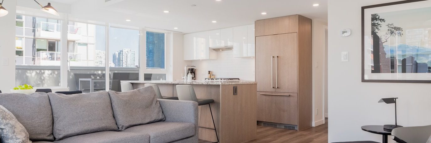 Modern open-concept kitchen and living room with light wood floors, a grey sofa, a large window with city views, white cabinets, and a built-in refrigerator. Framed art and minimalist decor accent the space.