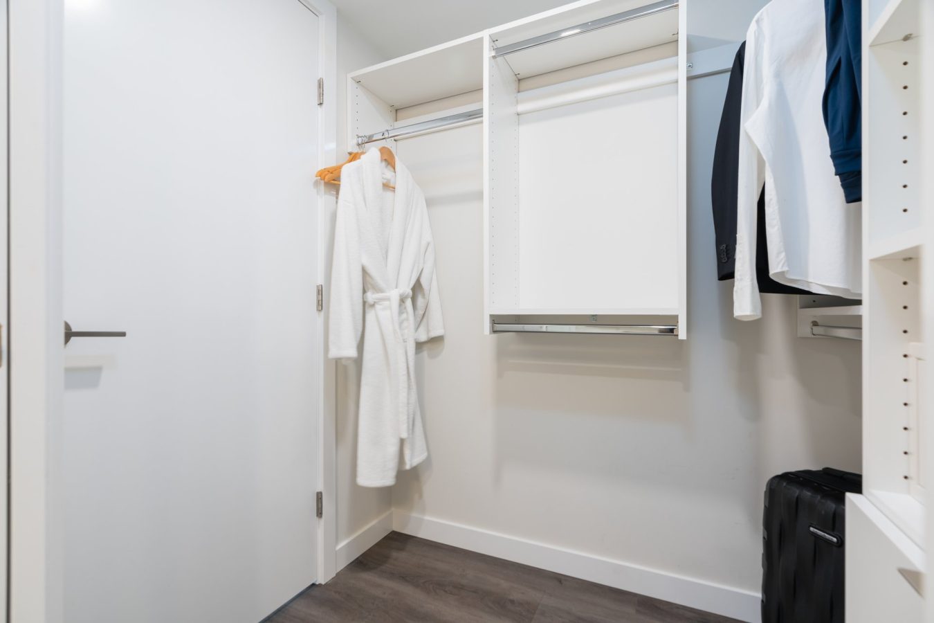 A modern walk-in closet with white shelves and hanging rods. A white bathrobe hangs on a wooden hanger, and a few shirts and a dark jacket hang on the right. A black suitcase sits on the floor near the wall.