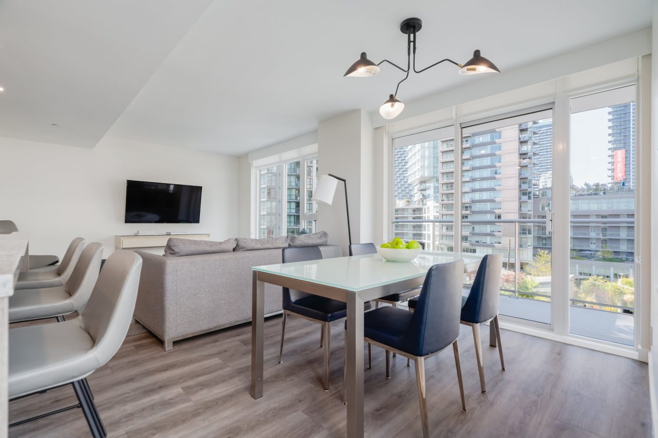 Modern open-plan living and dining area with large windows, gray sofa, wall-mounted TV, glass dining table with dark chairs, and a view of city buildings outside the balcony.