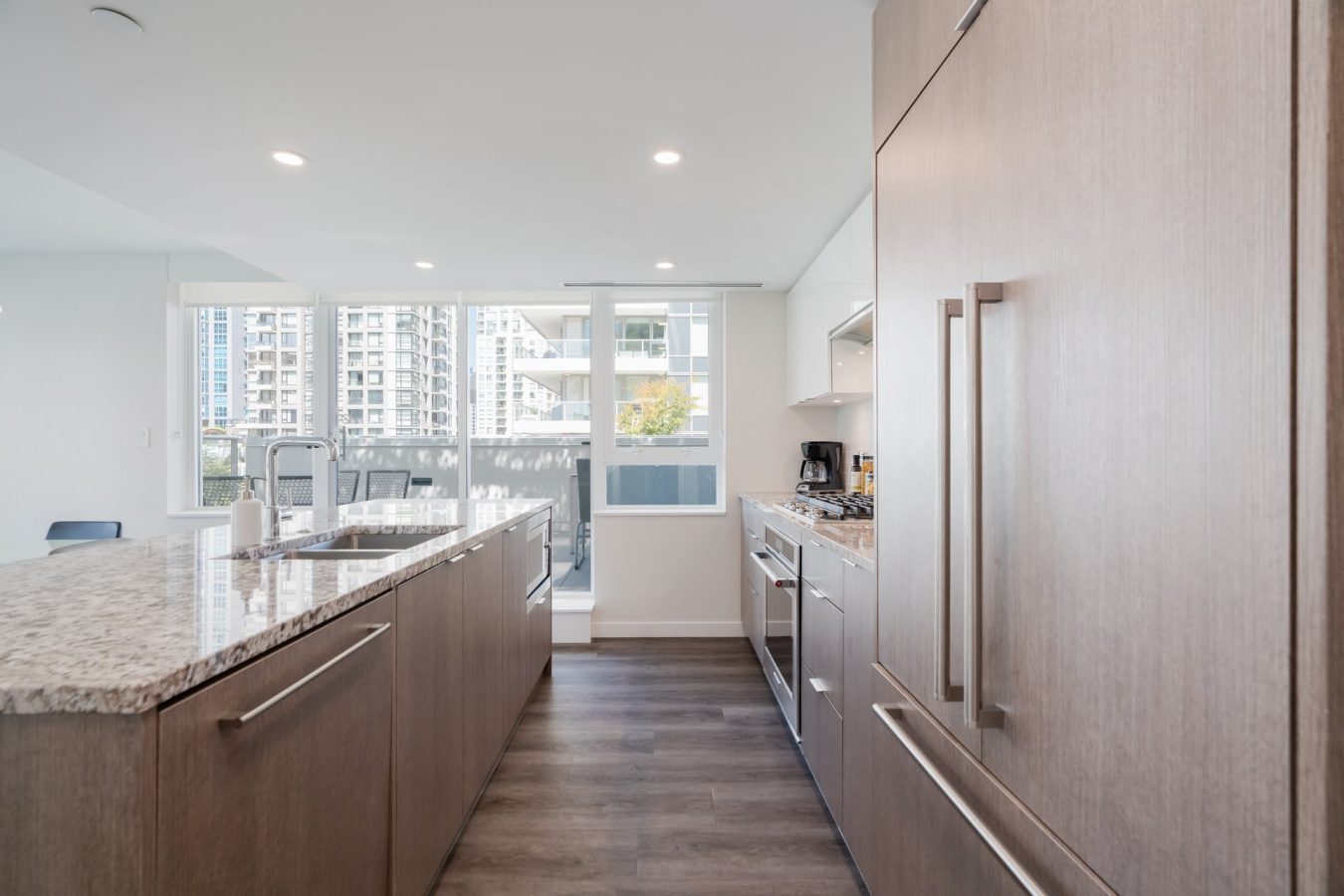 Modern kitchen with wood cabinets, a granite island, stainless steel appliances, and large windows letting in natural light; city buildings visible outside.