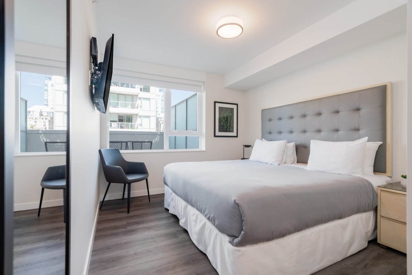 Modern, bright bedroom with a large bed, gray headboard, white bedding, two black chairs, wall-mounted TV, nightstands, large window, and cityscape view outside. Walls are white, and floors are dark wood.
