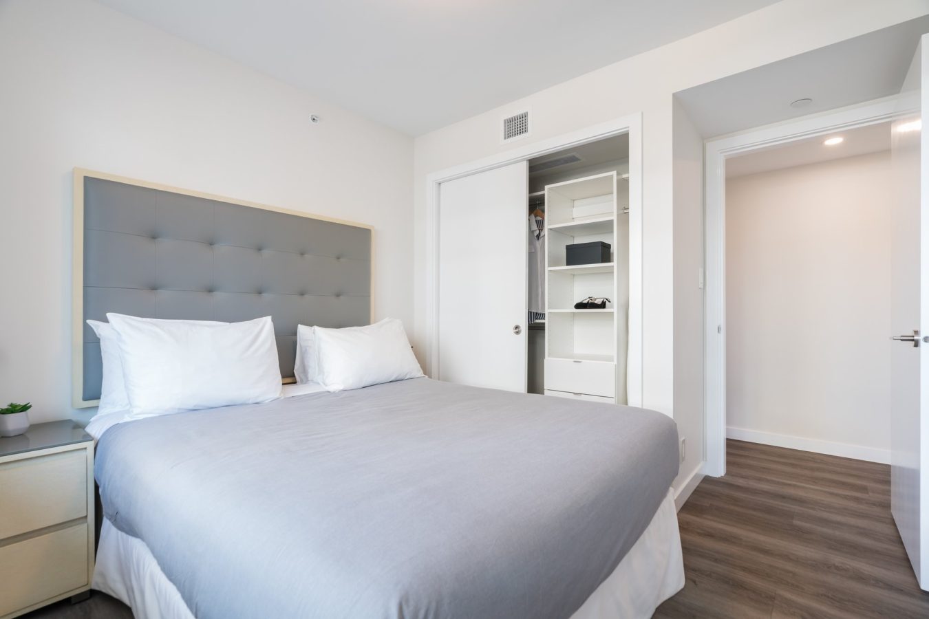 A modern bedroom with a neatly made bed with gray bedding, two white pillows, a padded headboard, a nightstand, an open closet with shelves, and a door leading to a hallway with wood flooring.