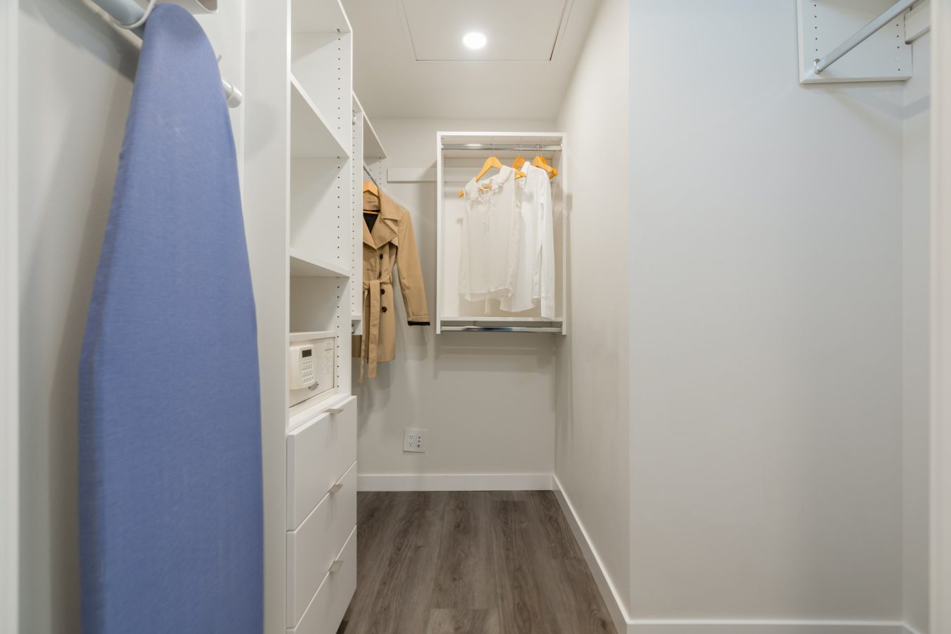 A modern walk-in closet with white shelving, a blue ironing board, hanging beige coats, white shirts on hangers, and dark wood flooring under bright ceiling lighting.