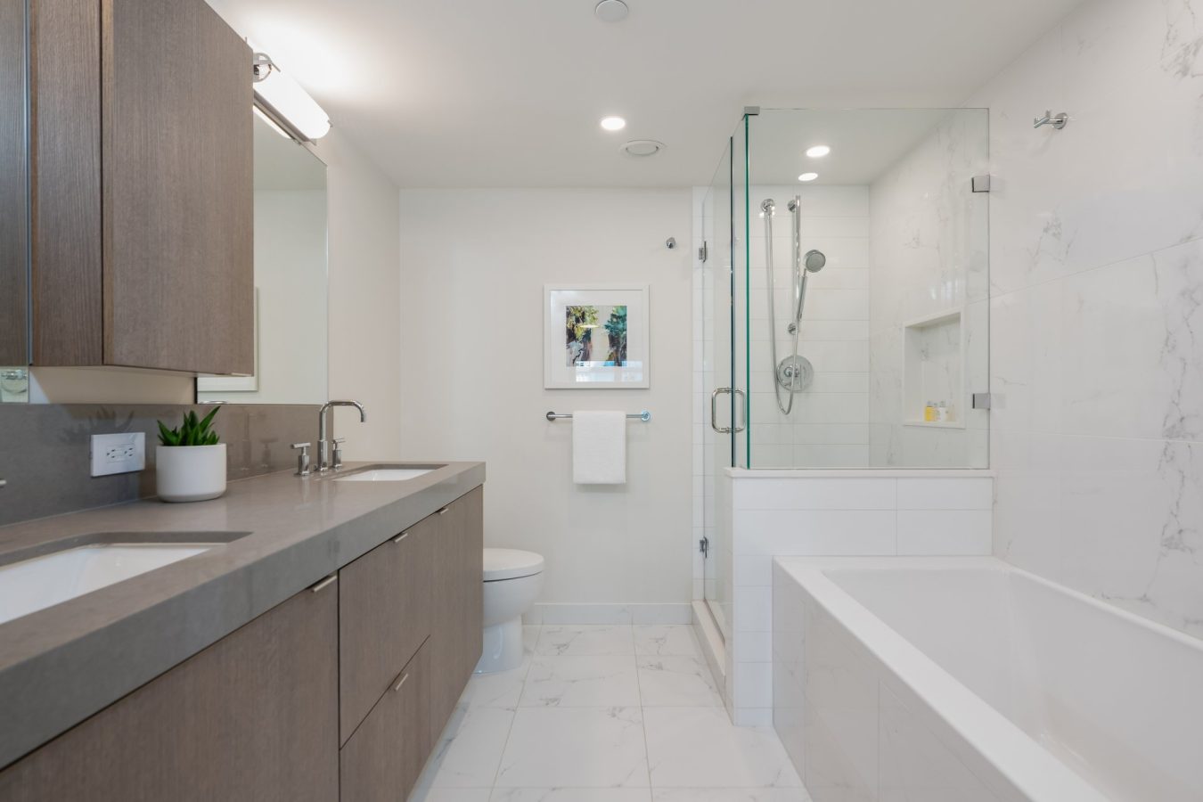 Modern bathroom with double sinks, wooden cabinets, a large mirror, a toilet, glass-enclosed shower, white bathtub, marble tile floor, and a small plant on the countertop. Walls are white and decor is minimal.