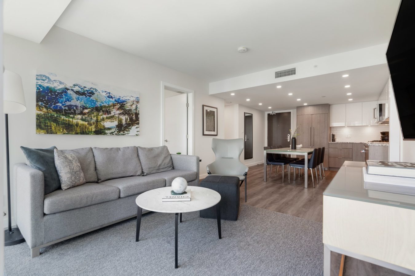 Modern living room with a gray sofa, armchair, round coffee table, and large wall art. Open layout leads to a dining area and contemporary kitchen with wood cabinets and recessed lighting.