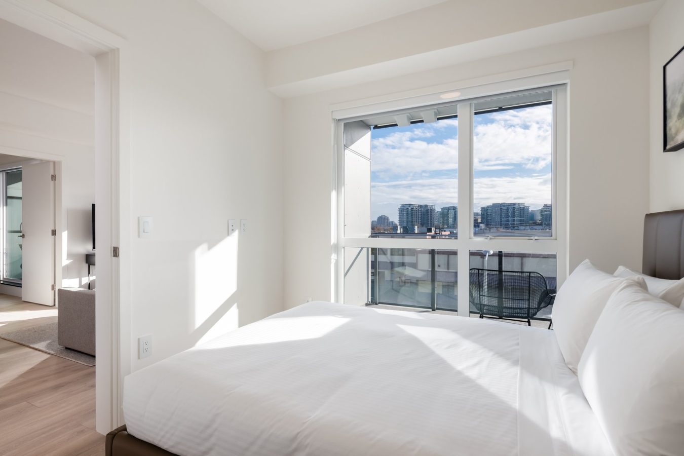 A bright, modern bedroom with a large window overlooking a cityscape. The bed has white linens, and there is a balcony with two chairs outside. Sunlight fills the room, creating a clean and inviting atmosphere.