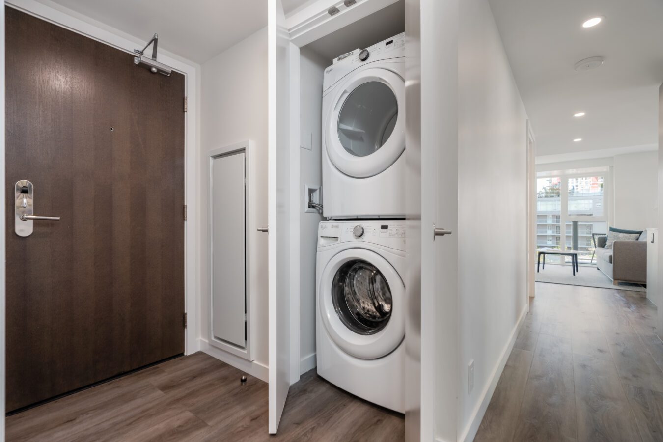 A stacked washer and dryer are inside a closet in a modern apartment hallway with wood flooring; a dark front door and a bright living area with large windows are visible.